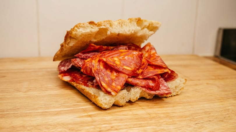 A rustic sandwich with a generous filling of sliced chorizo and salami on a wooden board.
