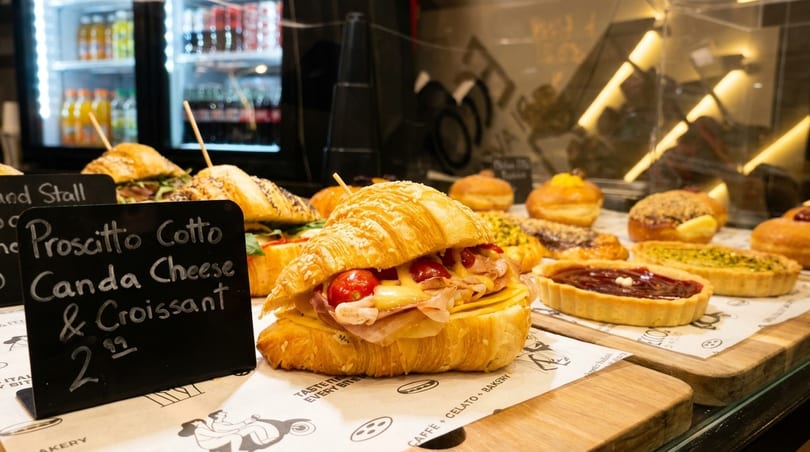 A croissant sandwich with prosciutto, cheese, and tomatoes, alongside pastries and a price sign.