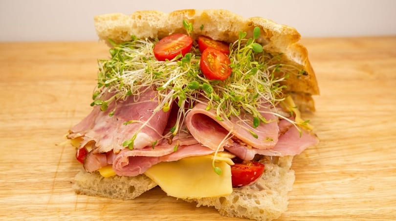 A close-up of a sandwich filled with ham, cheese, cherry tomatoes, and sprouts on a wooden board.