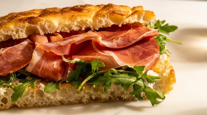 Close-up of a focaccia sandwich filled with prosciutto, arugula, and creamy white cheese.