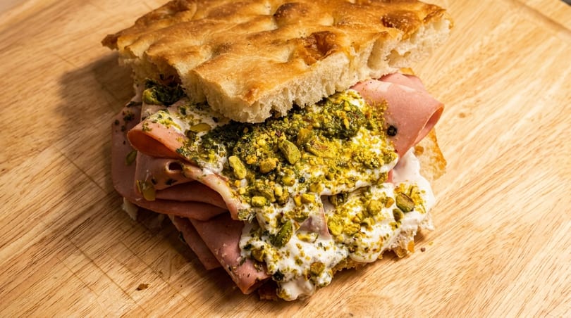 Mortadella sandwich on focaccia bread with pistachio pesto and burrata cheese on a wooden board.